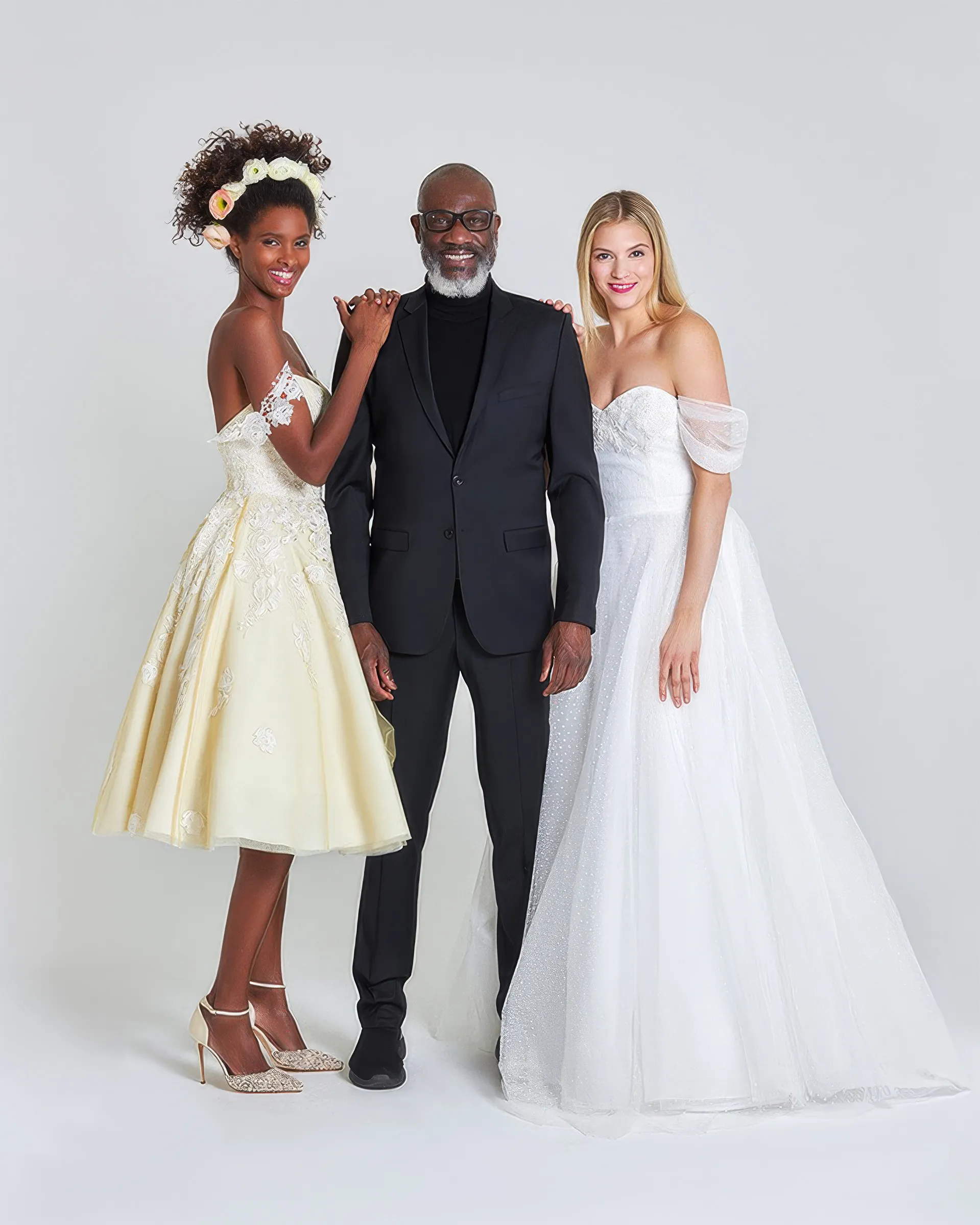 Yemi Osunkoya, in a black suit and polo neck, between two models: on the left Amber, a champagne tea-length tulle ballgown with lace appliqués and off-the-shoulder lace straps; on the right Toke, with off-the-shoulder tulle sleeves and a dotted tulle ballgown skirt.