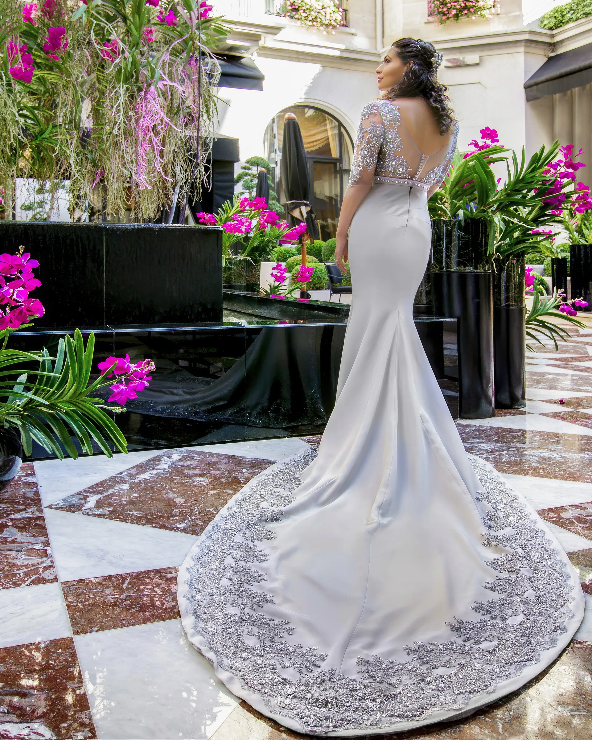 Erato, back, in the hotel courtyard: a deep V-back of illusion tulle and beaded lace above the fitted crepe skirt and its train bordered with silver appliqués.