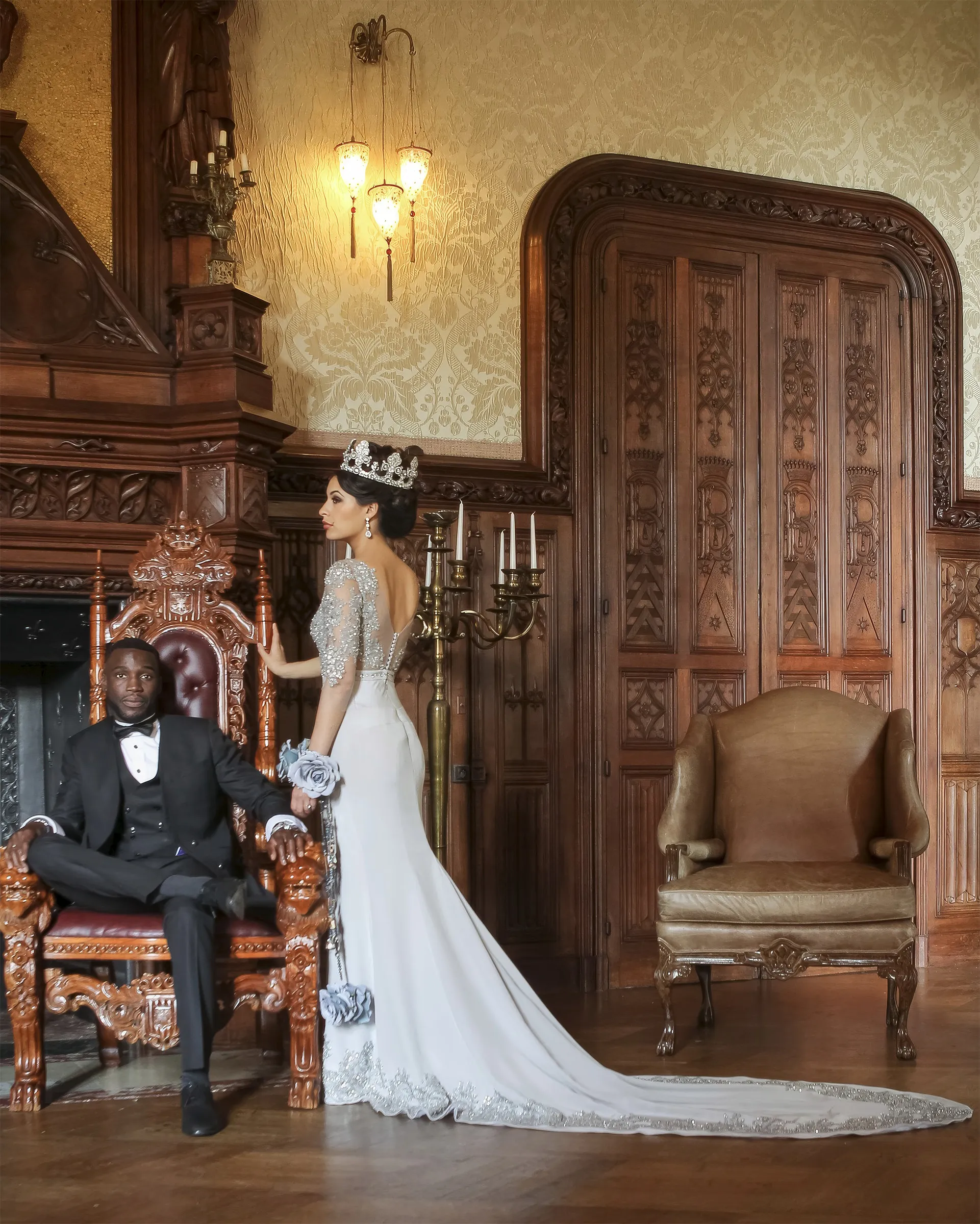 Erato beside a man in black tie seated on a carved throne, in a wood-panelled room: the beaded illusion back and the long appliquéd train.