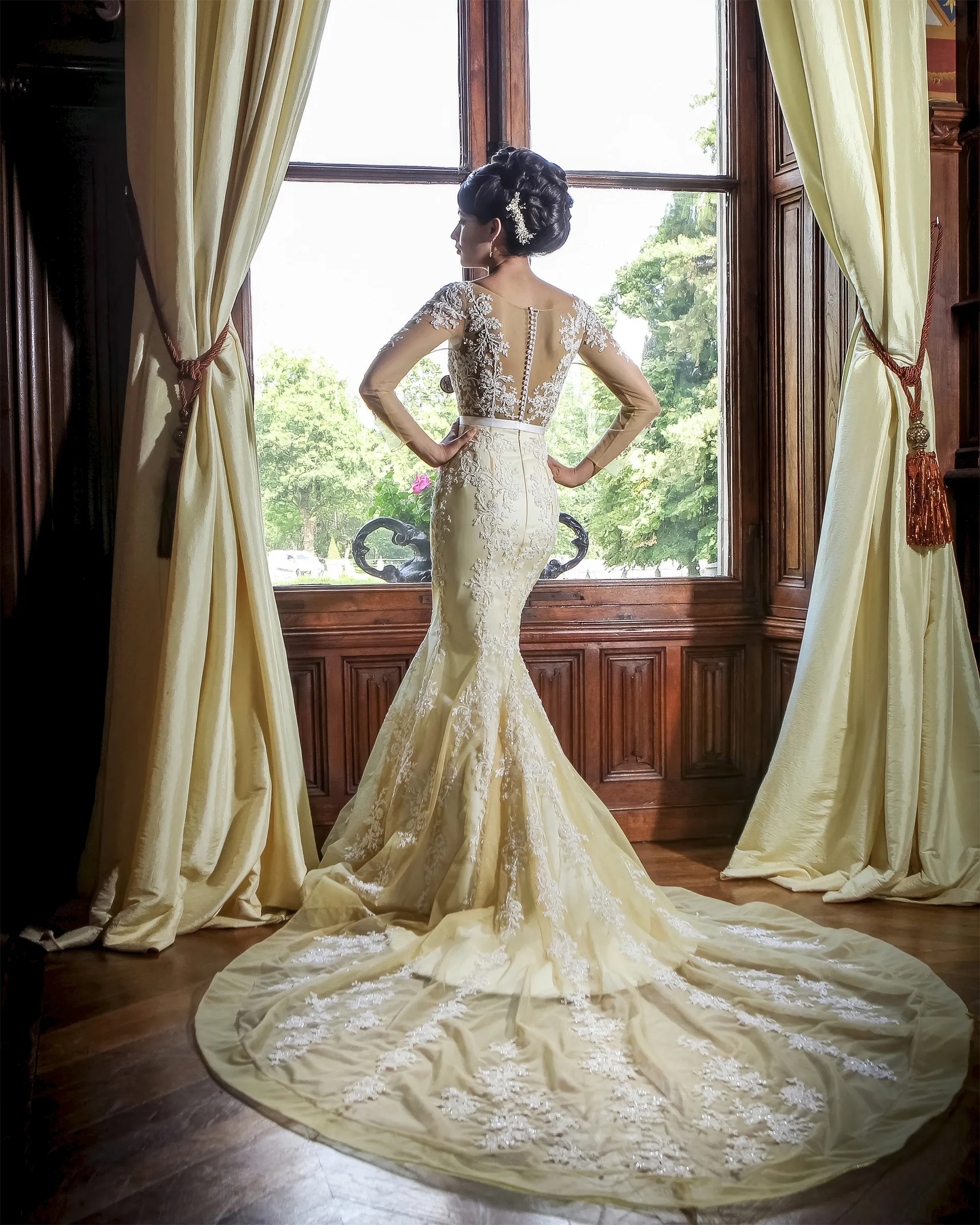 Euterpe, back, at a tall window between gold curtains: covered buttons down the illusion back and the trumpet skirt pooling into a lace-edged train.