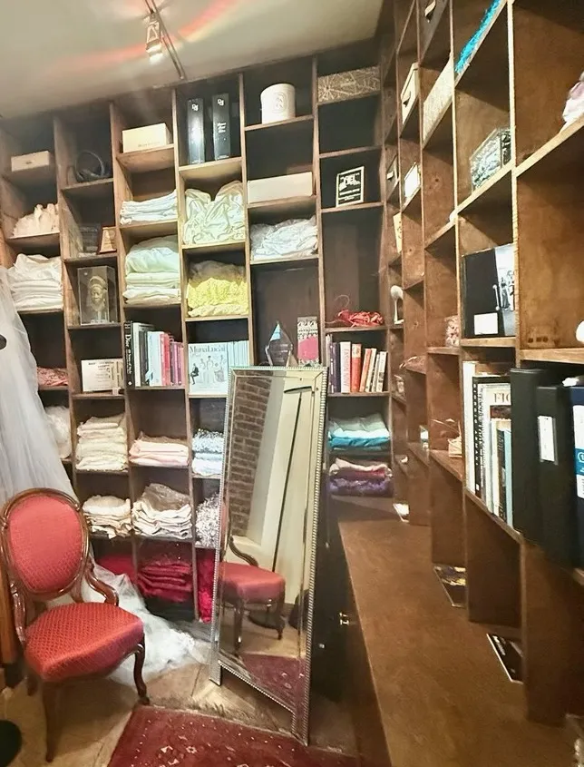 The changing room: wooden shelves of folded fabrics, books and accessories, a red velvet chair and a tall mirror.