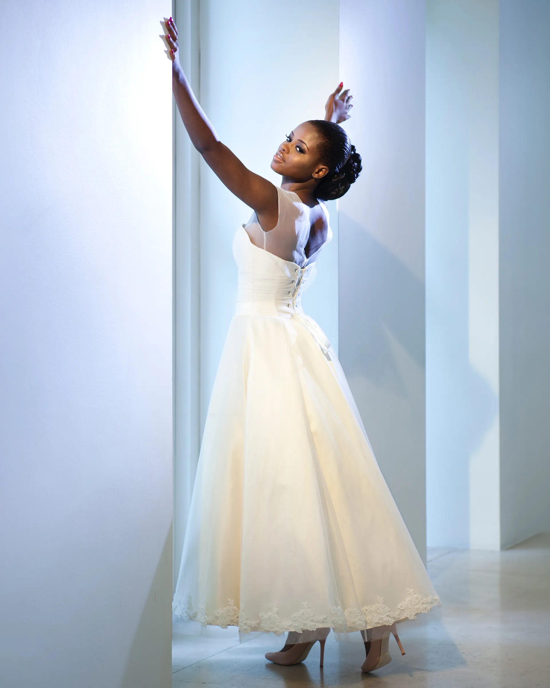 Giselle, arms raised to the wall: the corseted bodice laces up the back beneath the sheer chemise, and the tea-length skirt falls full to a lace hem.