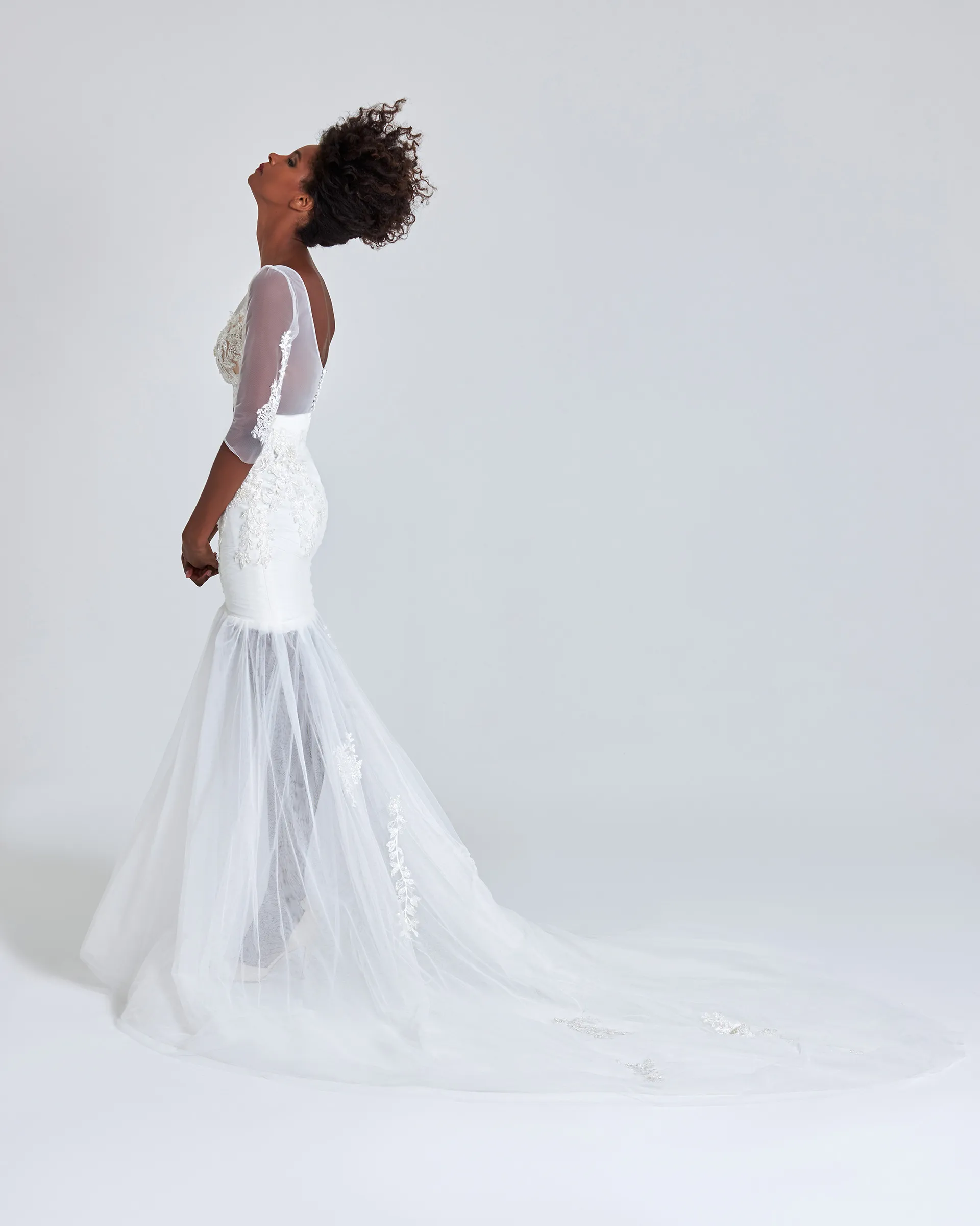 Ife, side, head tipped back: lace appliqués trail down the sheer sleeve, and the layered tulle mermaid skirt spreads into a long train.