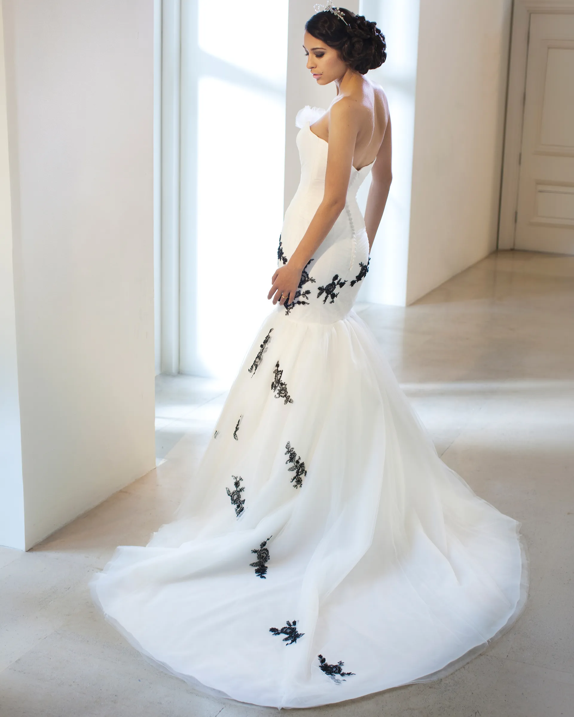 Odile from behind in a white room: black appliqués dot the tulle fishtail as it spreads into a chapel train, with covered buttons down the back.