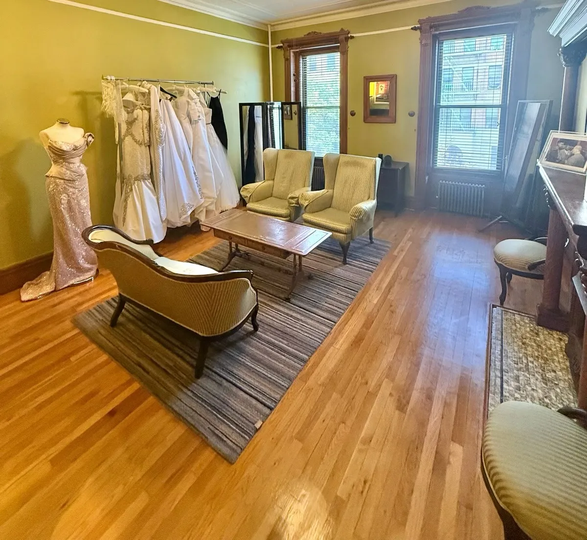 The Kosibah salon: striped wingback chairs and a settee around a low table, a couture gown on a dress form and bridal gowns on a rail, tall windows behind.