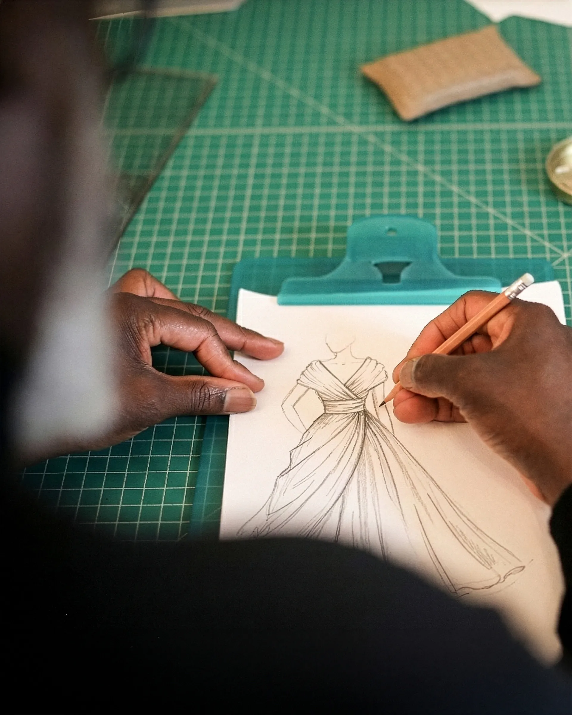 Yemi’s hands drawing a draped, off-the-shoulder gown in pencil on a clipboard, on the green cutting mat.