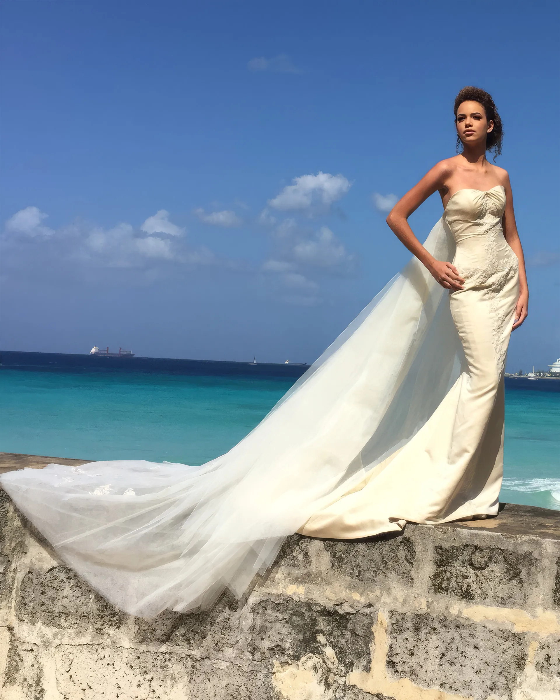 Thalia standing on a stone sea wall above turquoise water, the detachable tulle Watteau train streaming behind the fitted satin sheath.