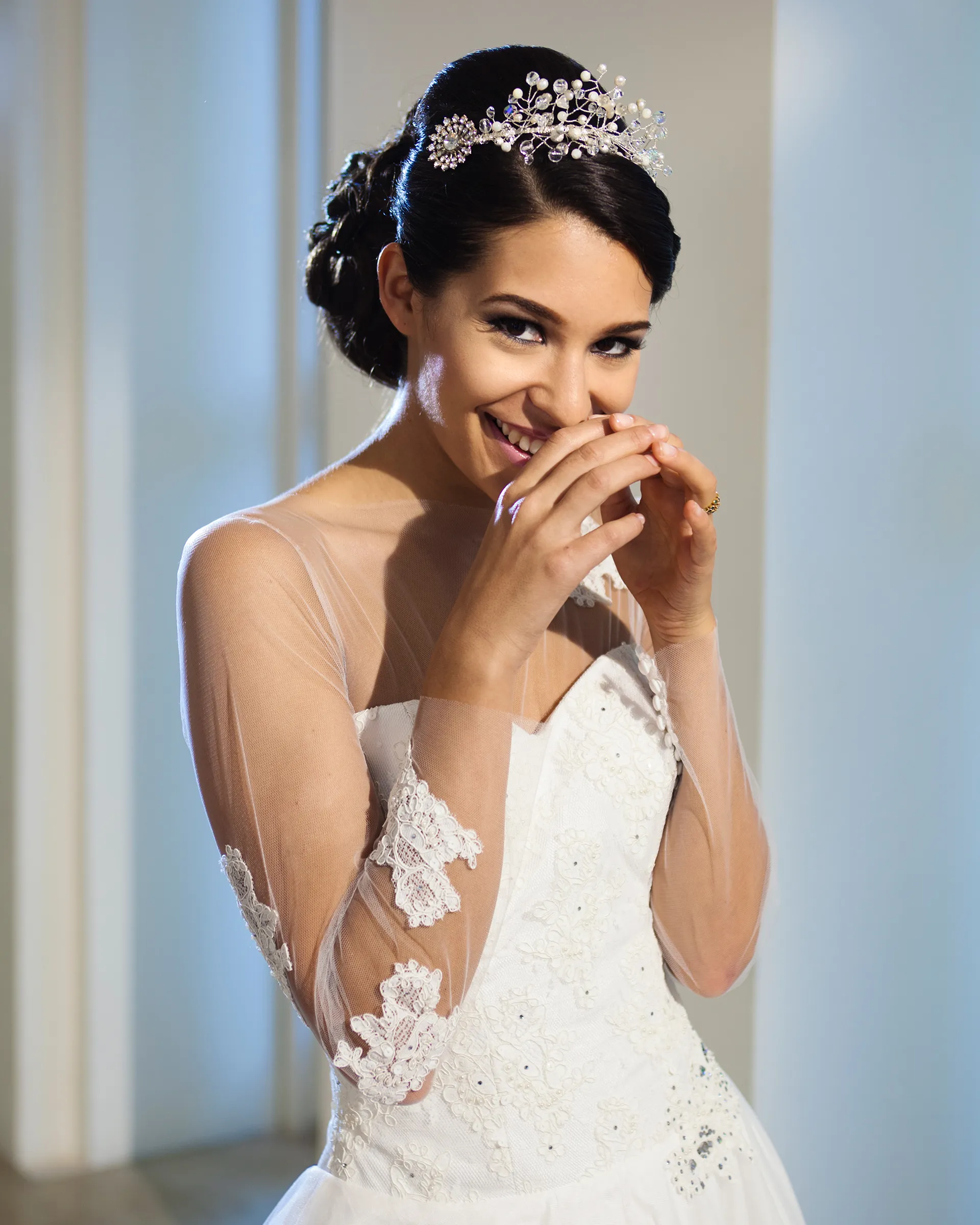 Titania, front detail, laughing with her hands to her face: lace appliqués trail down the sheer tulle sleeves over a beaded lace sweetheart bodice.