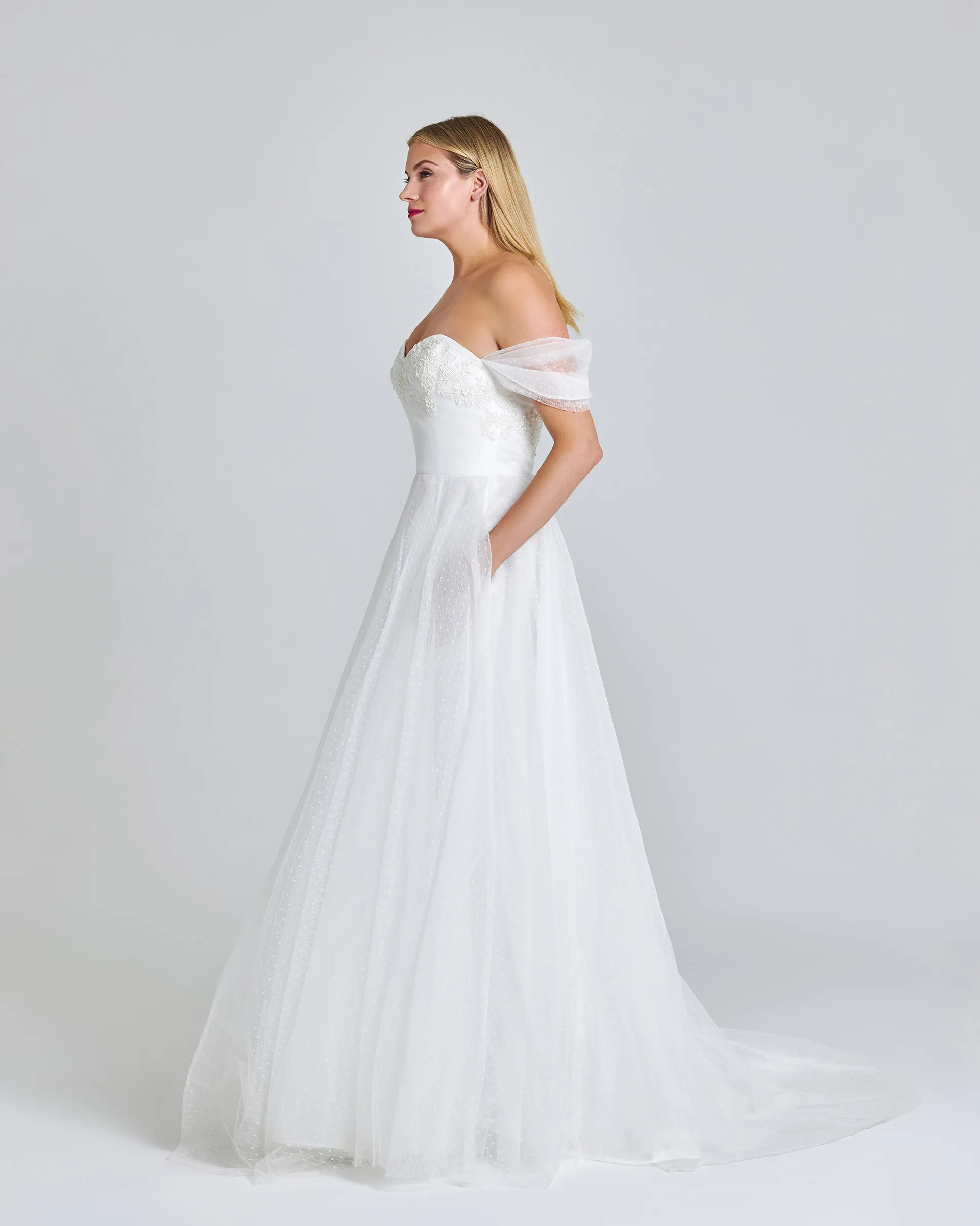 Toke, side, hand in a pocket of the ballgown skirt, which falls in soft layers of dotted tulle to a short train.
